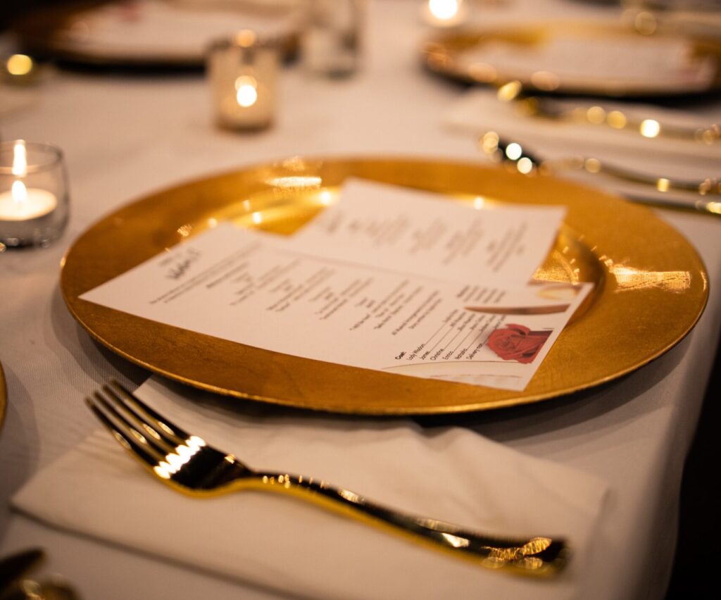 Fundraising gala dinner setting of a gold plate with a menu, fork and napkin, and candlelight